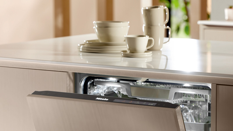 Teilgeöffnete Miele Spülmaschine in heller Küche mit Geschirr auf der Arbeitsplatte darüber.